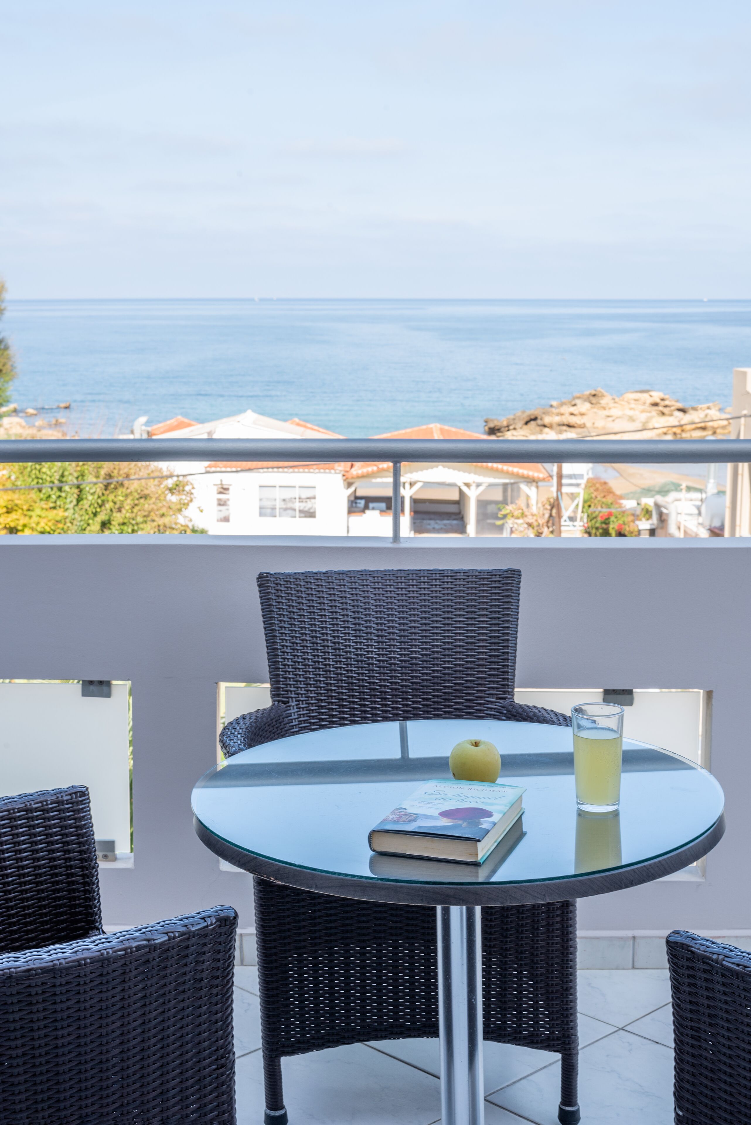 Una mesa redonda de vidrio con sillas de mimbre en un balcón con vista al mar.
