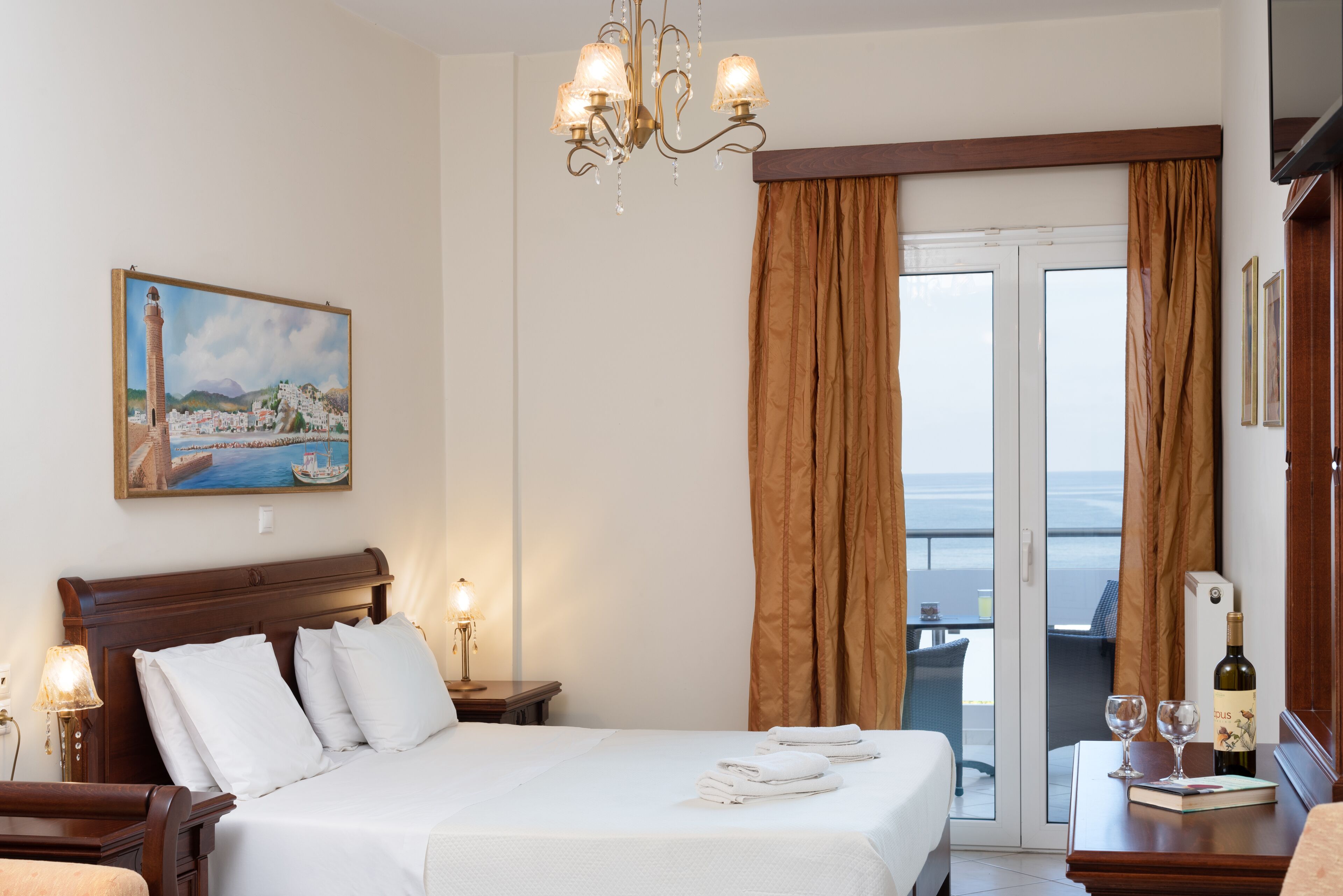 A hotel room with a bed, a painting of a coastal scene, a chandelier, and a view through the curtains.