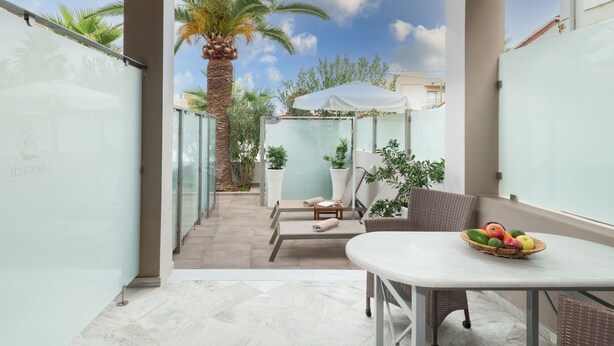 Un patio moderno al aire libre con pared de vidrio, una mesa blanca con frutero, sillas de mimbre y un paraguas blanco.