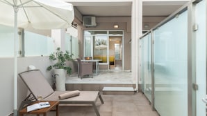 Un área de lounge al aire libre, moderna, con pared de vidrio, zona de estar con mesa y sillas, y una planta en maceta.