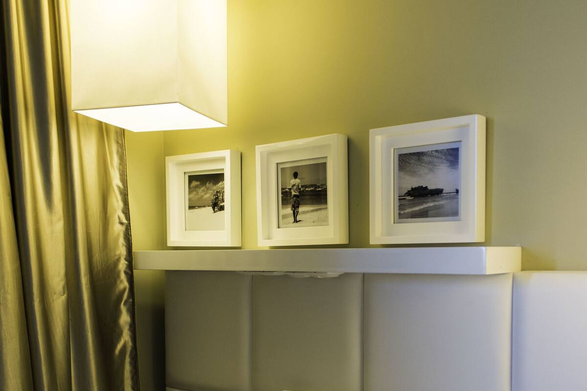 A hotel room with a bed, a headboard, a shelf with framed pictures, and a hanging light fixture.