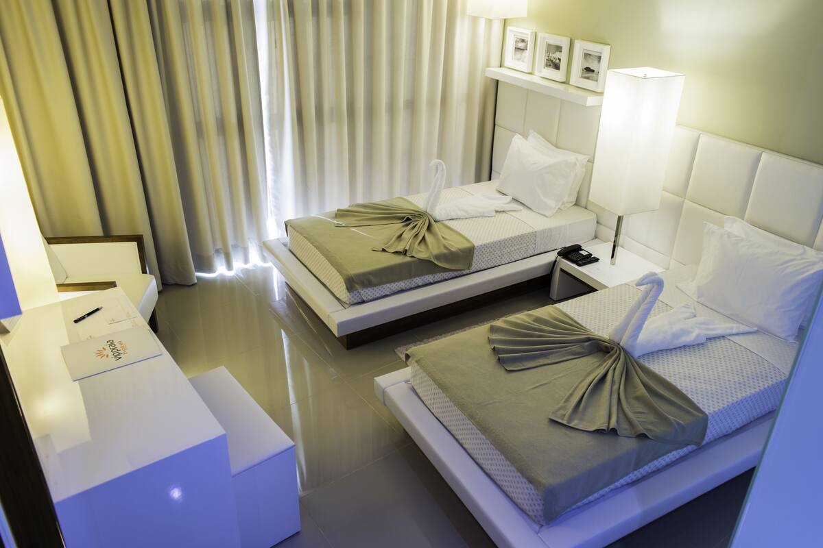 A hotel room with two beds, a bedside table, a lamp, and a window with curtains.