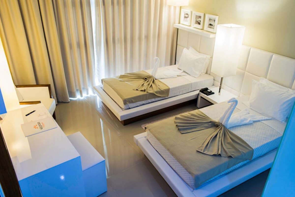 Una habitación de hotel con dos camas, una mesita de noche, una lámpara y un ventanal con cortinas.