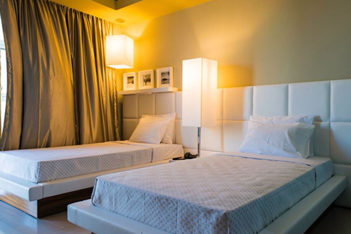 Habitación de hotel con dos camas, un ventanal con cortinas y lámparas fijadas a la pared.