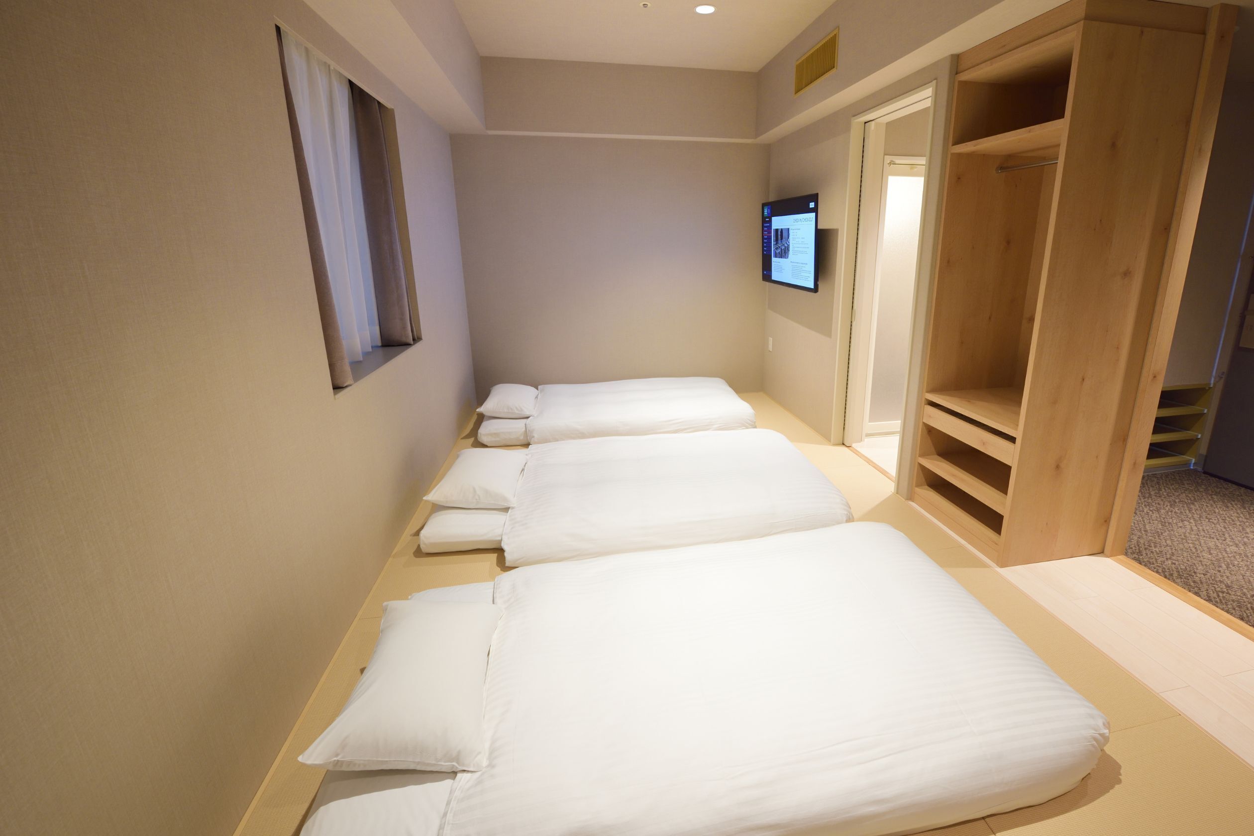 A hotel room with a long, narrow layout featuring a series of beds aligned along one wall, a wooden wardrobe, and a television mounted on the wall.