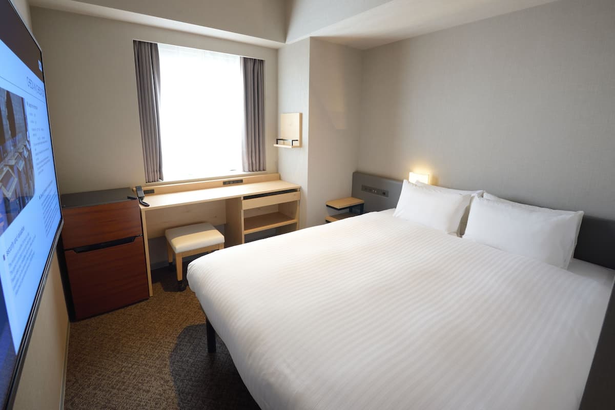 Une chambre d’hôtel avec un grand lit, un bureau avec une chaise, une petite table de chevet et un téléviseur placé sur le côté gauche.
