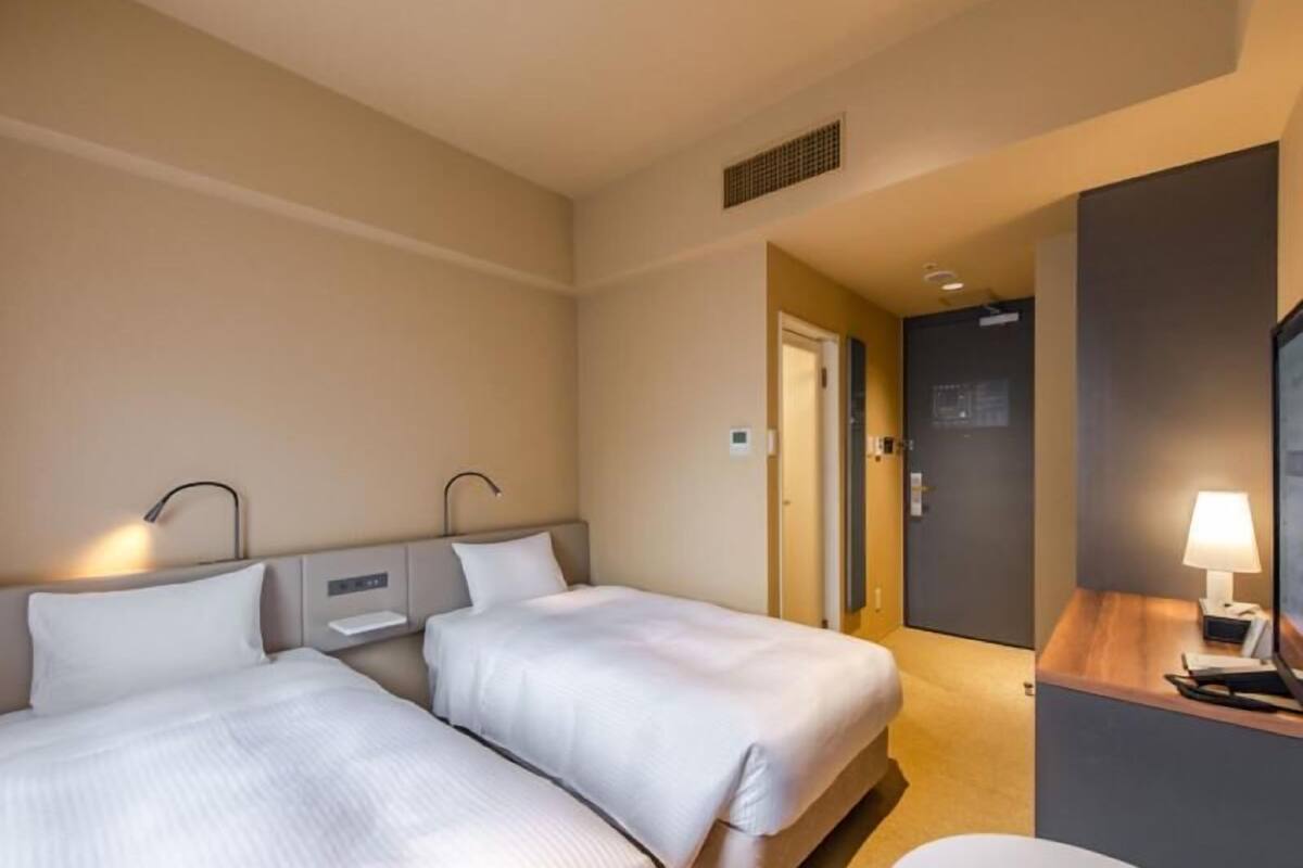 Une chambre d’hôtel avec deux lits, des tables de chevet, un bureau avec une lampe et une porte donnant sur une autre pièce.