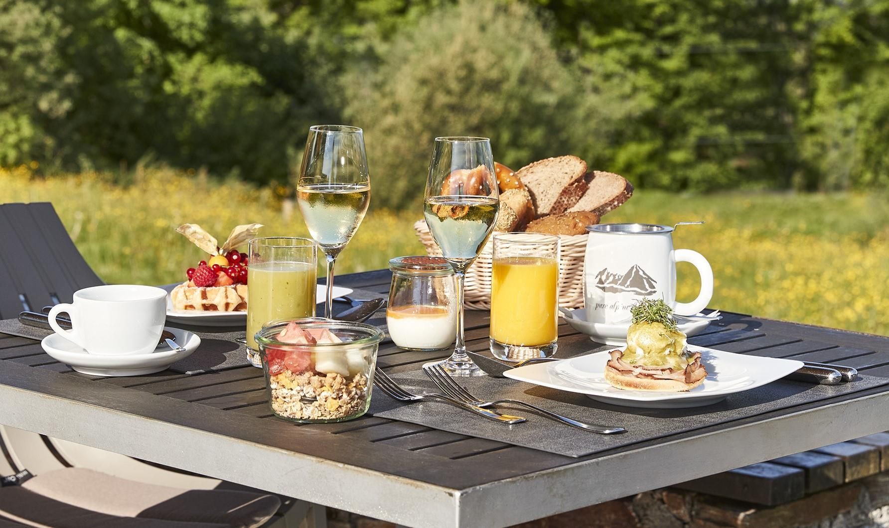 Una mesa al aire libre con diversos alimentos para el desayuno, incluyendo pan, una taza, un vaso de jugo, un vaso de agua y una copa de vino.