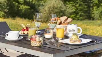 Una mesa al aire libre con diversos alimentos para el desayuno, incluyendo pan, una taza, un vaso de jugo, un vaso de agua y una copa de vino.