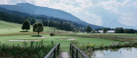 Un puente de madera que conduce a un campo de golf con un estanque, árboles y una montaña al fondo.