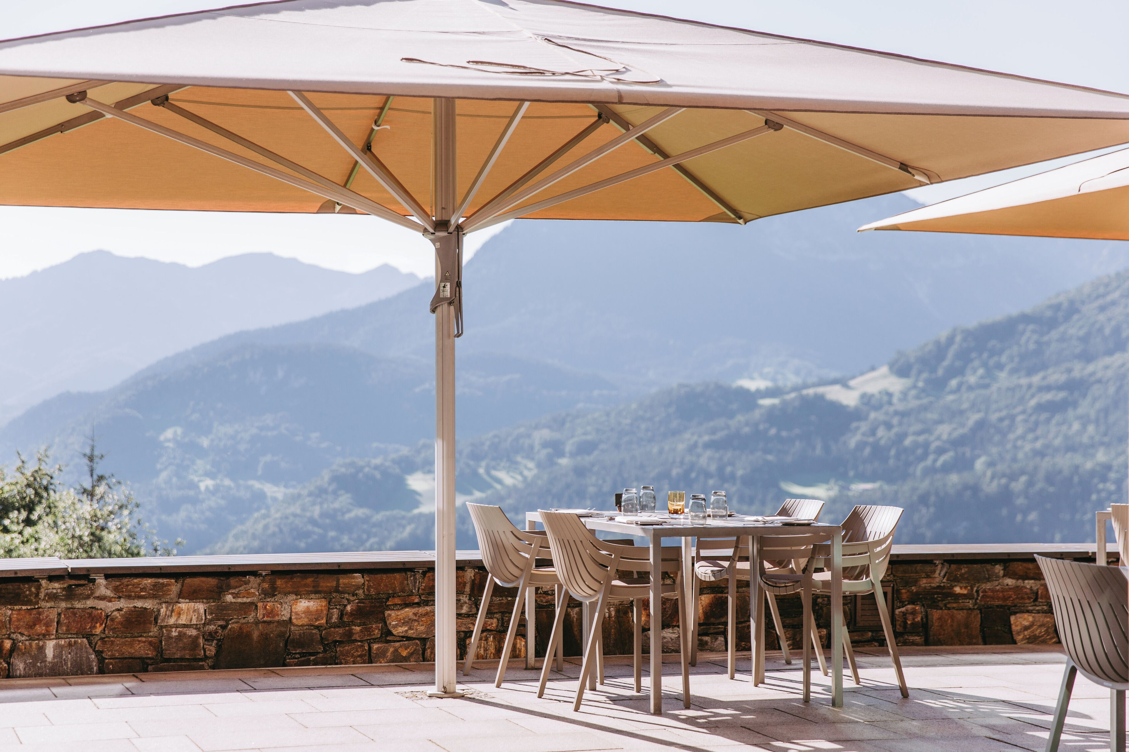Un área de comedor al aire libre con mesa y sillas bajo un paraguas grande, con vistas a un paisaje montañoso.