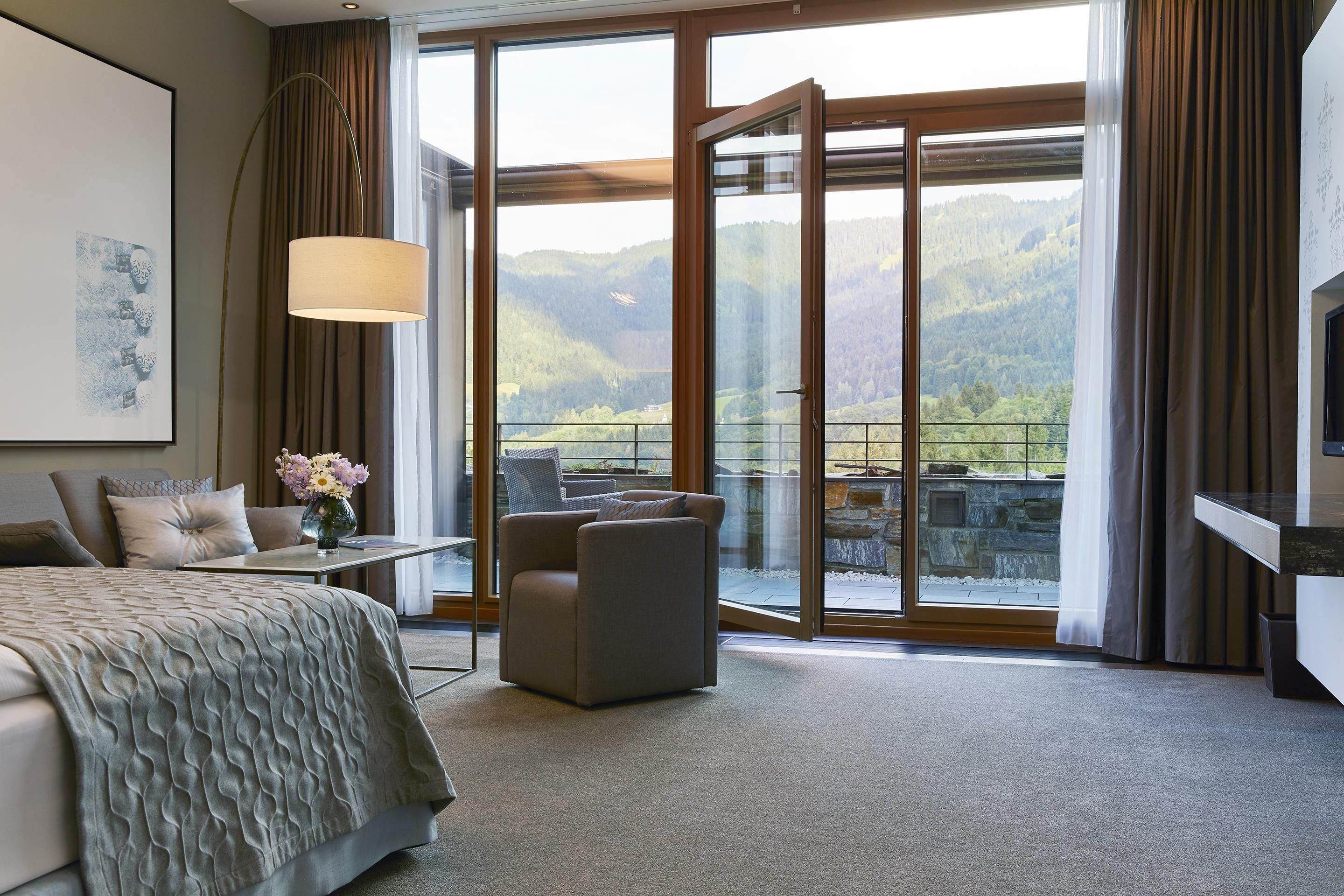 A hotel room with a bed, a desk, a chair, a large window with a mountain view, and a painting on the wall.