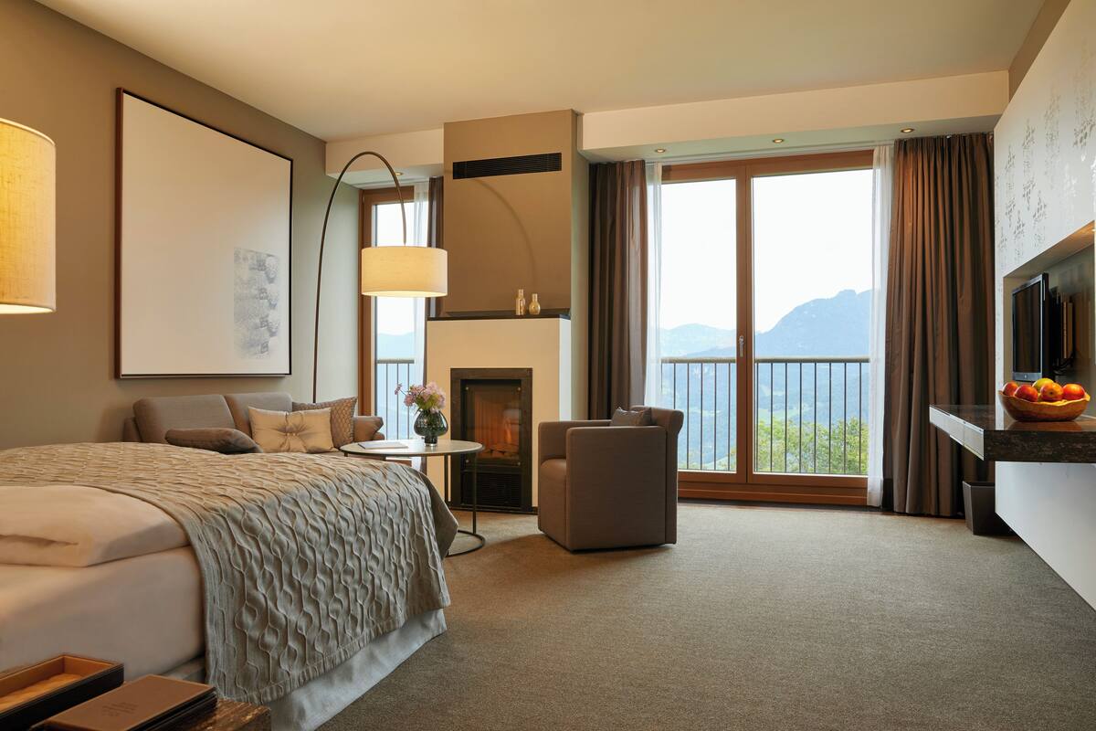 Ruime hotelkamer met een groot bed, zithoek, open haard en balkon met uitzicht op de bergen.