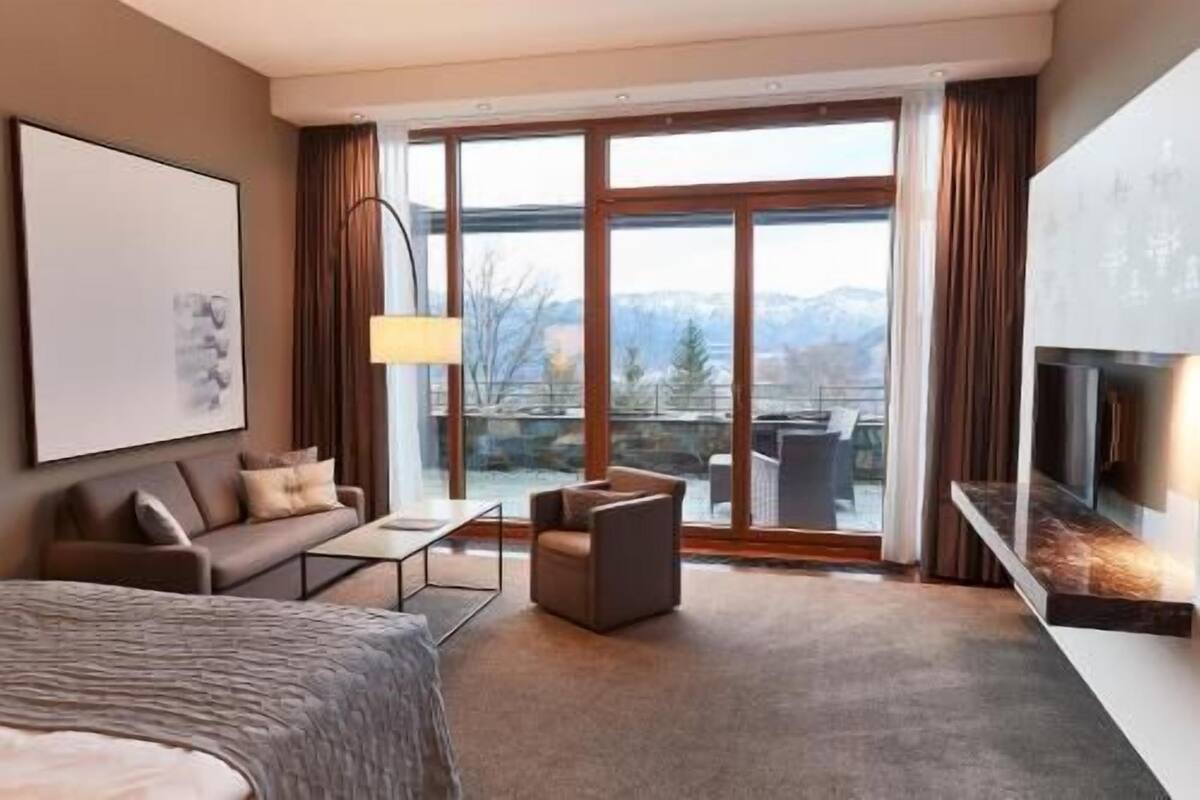 Een moderne hotelkamer met een groot bed, een bank, een kleine tafel, een televisie en een balkon met uitzicht op bergen.