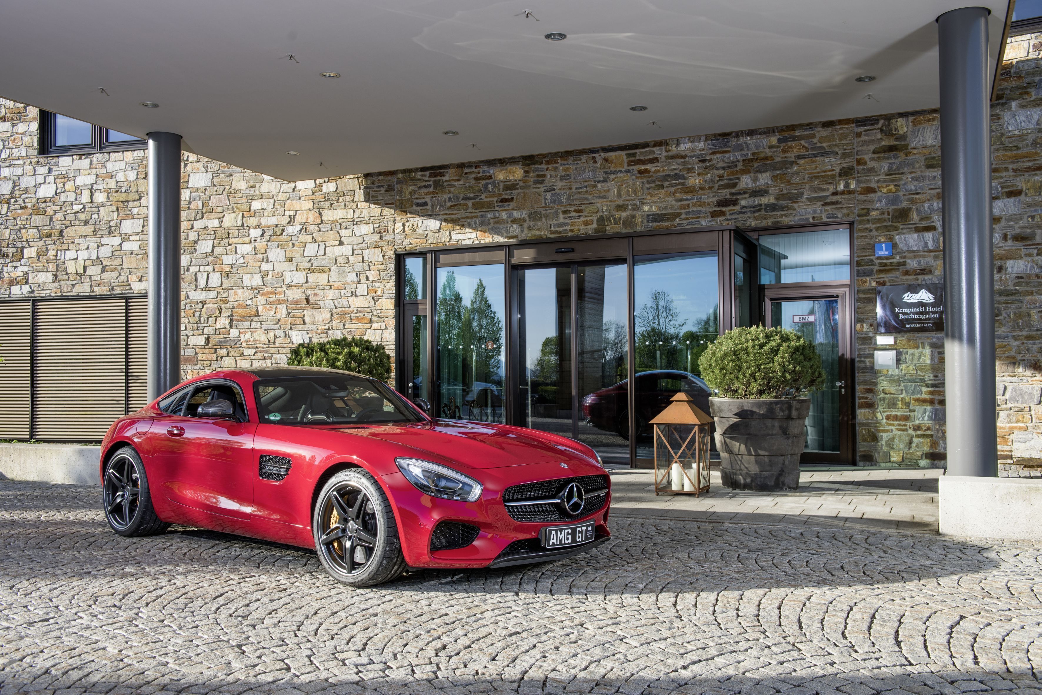 Une voiture de sport rouge est garée devant un hôtel aux murs en pierre et à l’entrée en verre.