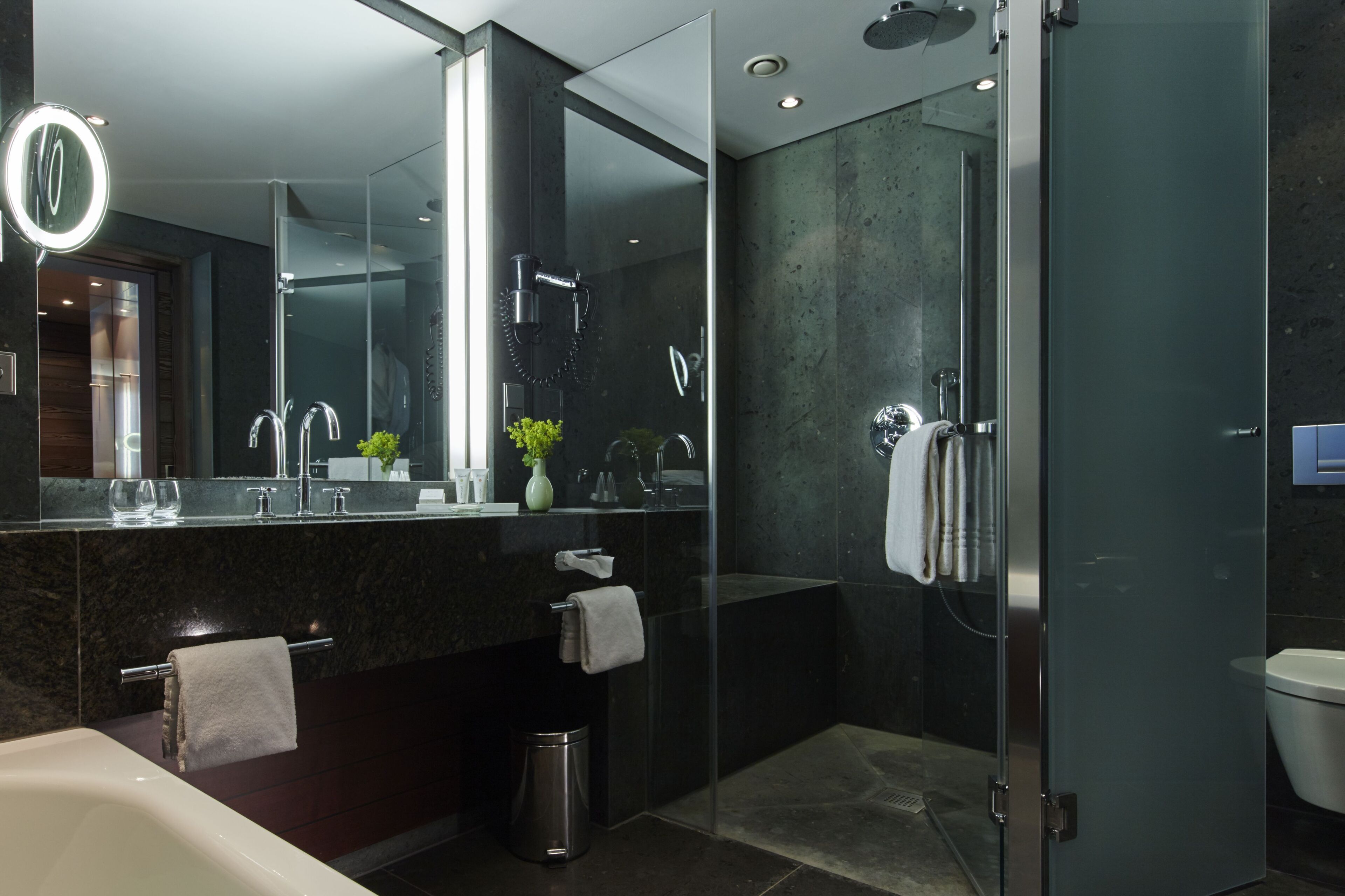 A modern bathroom with a glass-enclosed shower, a large mirror with a light, and a dark marble countertop.