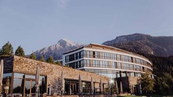 Un hotel moderno con fachada de piedra, ventanas grandes y mesas exteriores con sombrillas.
