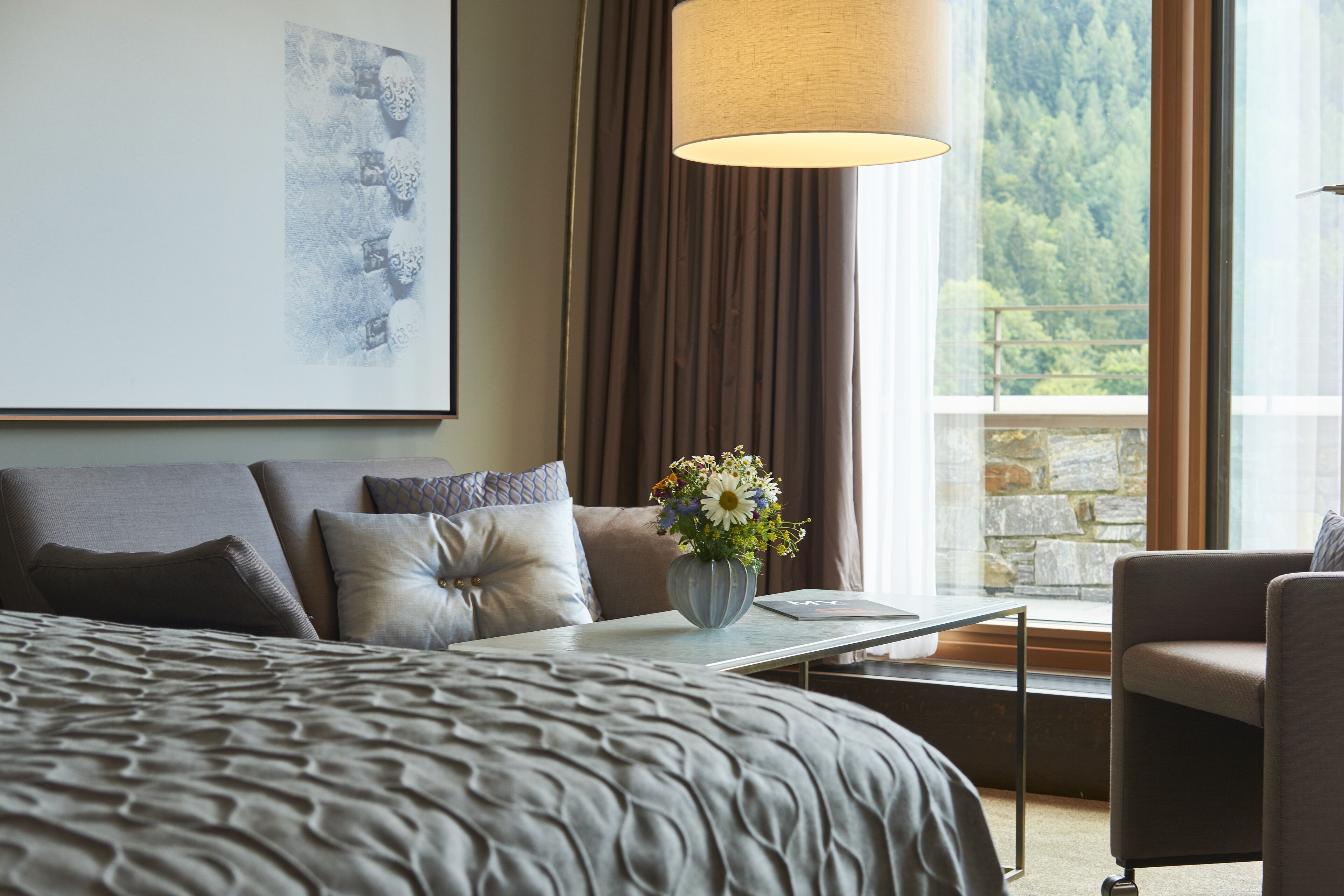 Een slaapkamer met een groot bed, een nachtkastje met een bloevas, een stoel en een raam met uitzicht op een bos.