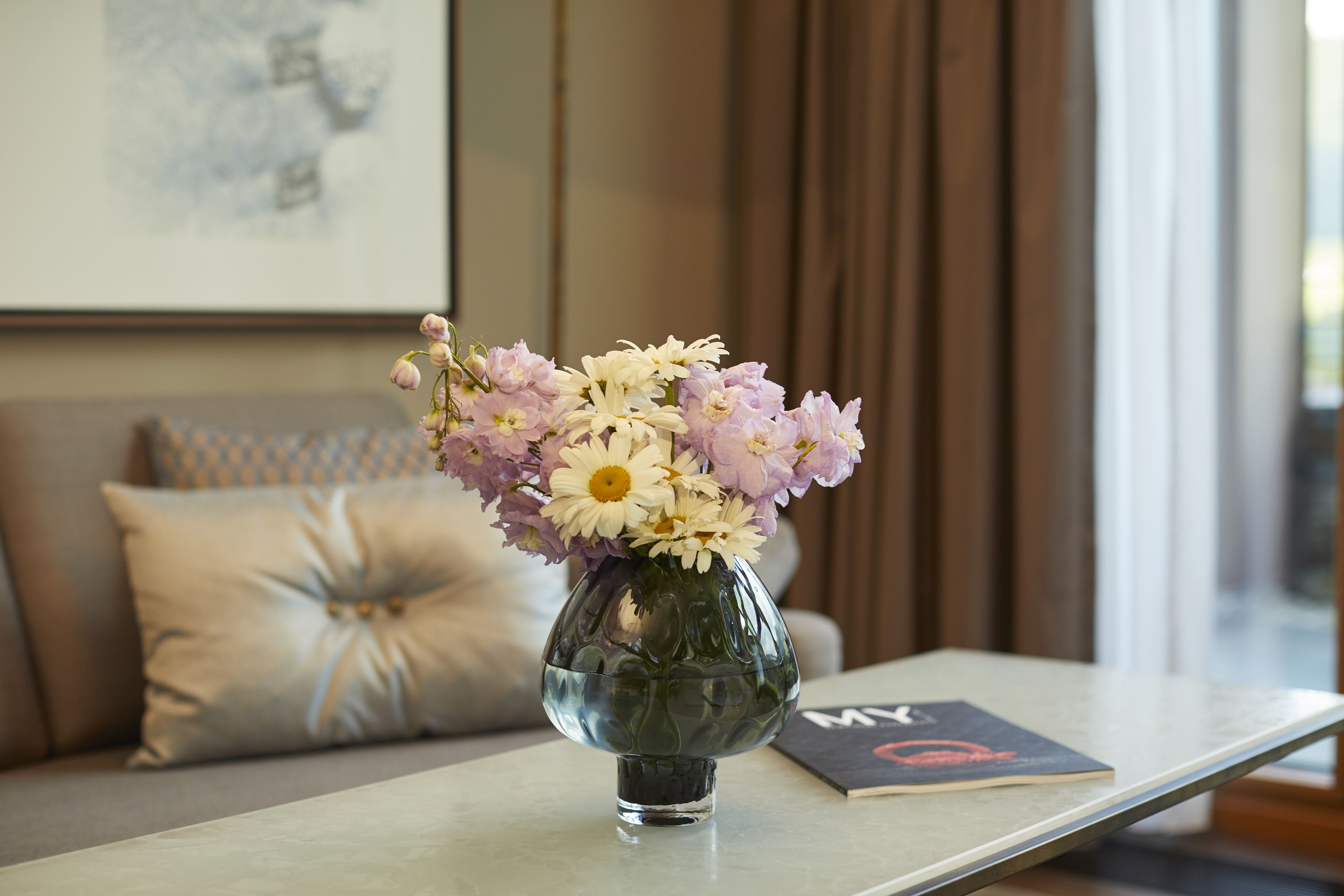 A vase with flowers on a table in a room with a couch and a framed picture on the wall.