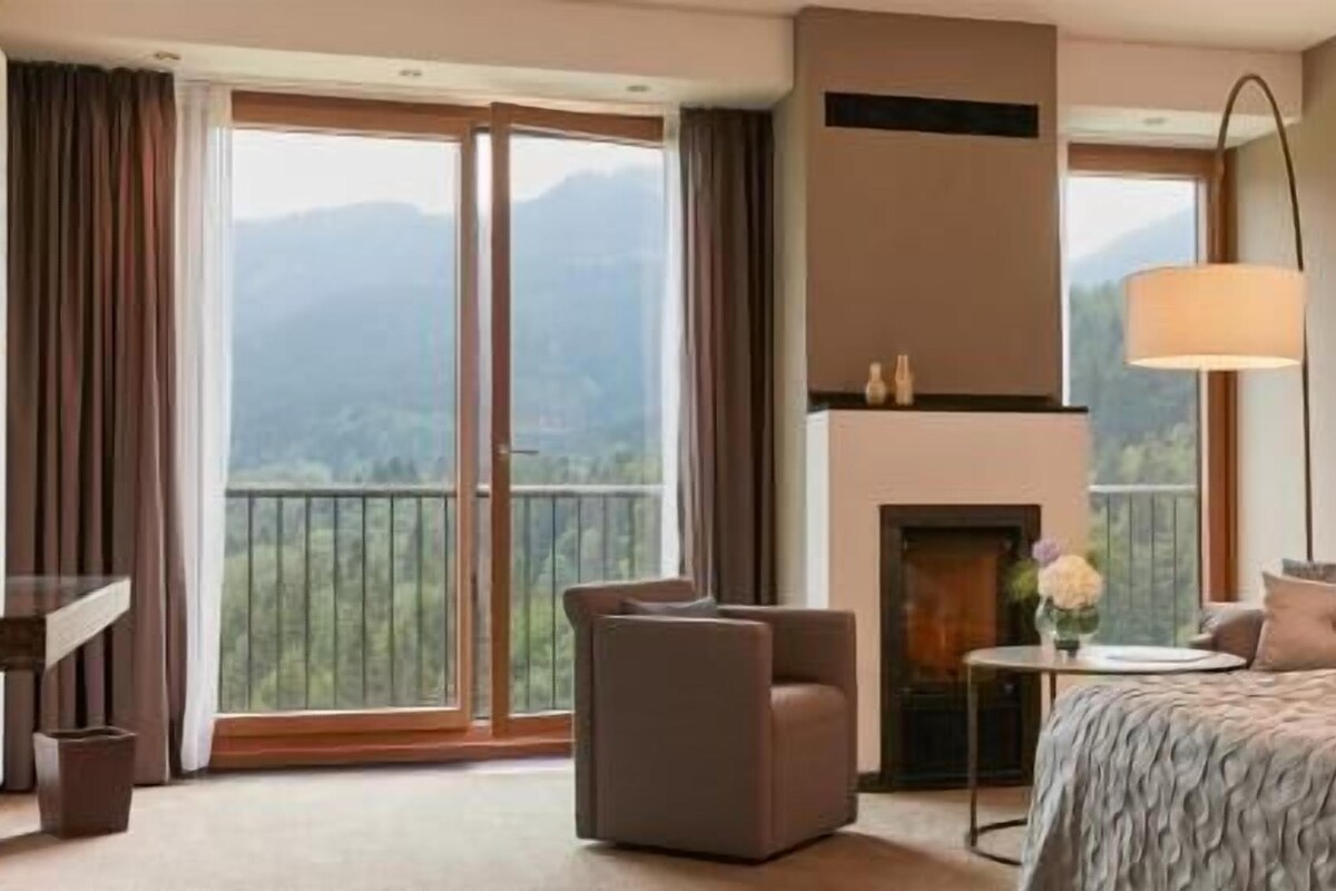 A hotel room with a large window, a fireplace, a sofa, and a small table.