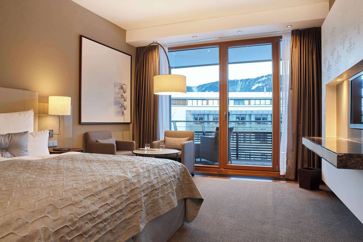 Hotelkamer met een groot bed, bureau, twee stoelen, televisie en balkon met uitzicht op gebouwen en heuvels.