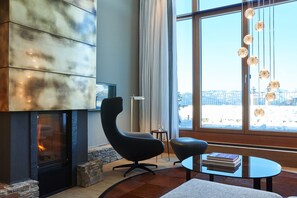 Een moderne woonkamer met een open haard, een comfortabele fauteuil, een tafeltje met boeken, en een groot raam dat uitzicht biedt op een besneeuwd landschap.