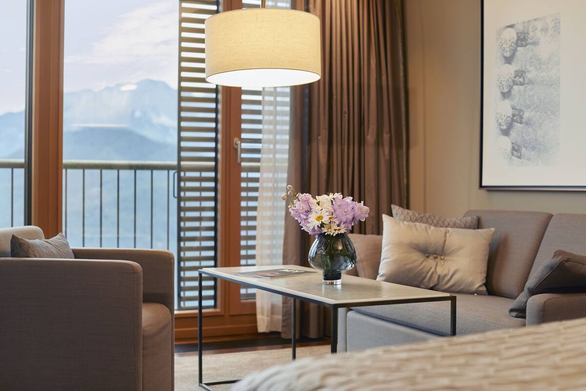 Een hotelkamer met een bank, een klein tafeltje met een bloevas, een lamp en een raam met uitzicht op bergen.