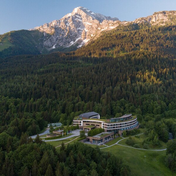 Un bâtiment moderne doté d’un toit incurvé, entouré d’une zone boisée et avec des montagnes en arrière-plan.