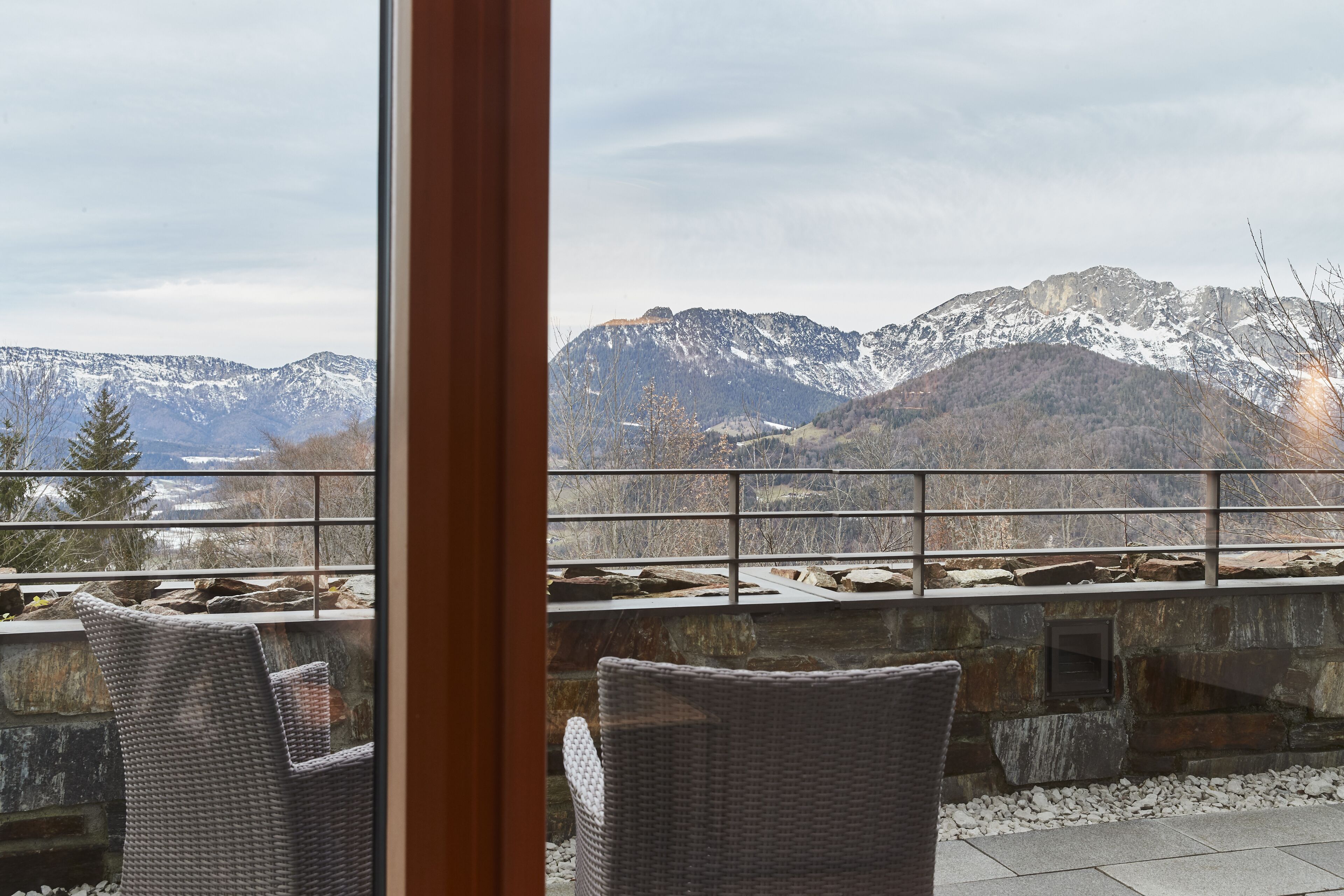 Vista da un balcone con sedie in vimini, che si affaccia su montagne innevate e un muro in pietra.