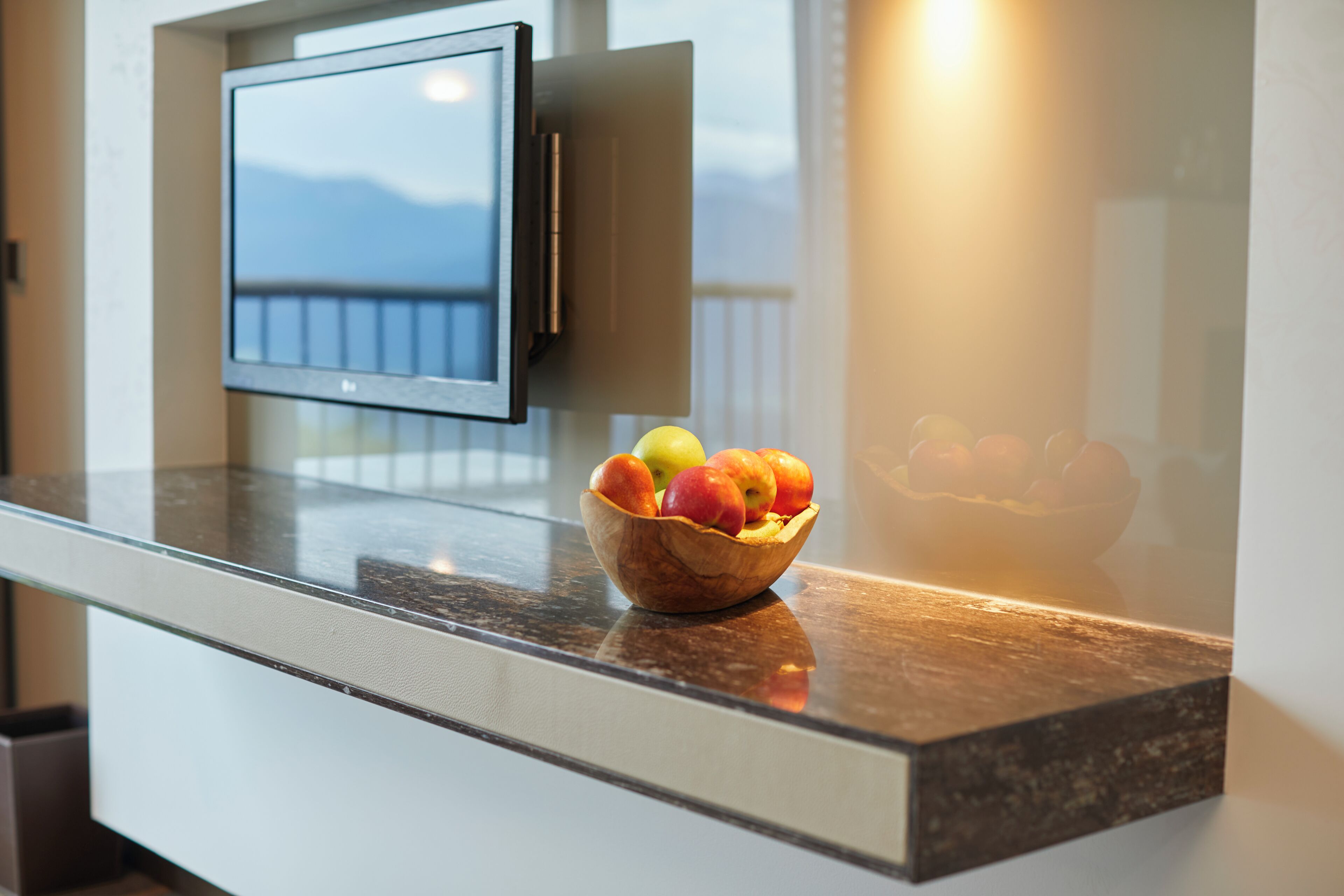 Una cucina moderna con una ciotola di legno piena di frutta su un piano di lavoro, una televisione a schermo piatto montata a parete e vista sulle montagne attraverso una finestra.