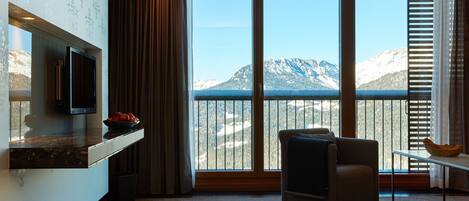 A modern hotel room with a large window offering a mountain view, a flat-screen TV mounted on the wall, and a comfortable seating area with a view.