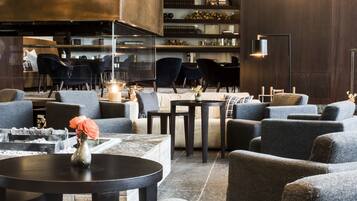 A modern lounge area with sofas, tables, and a fireplace. There are bookshelves and a coffee machine in the background.