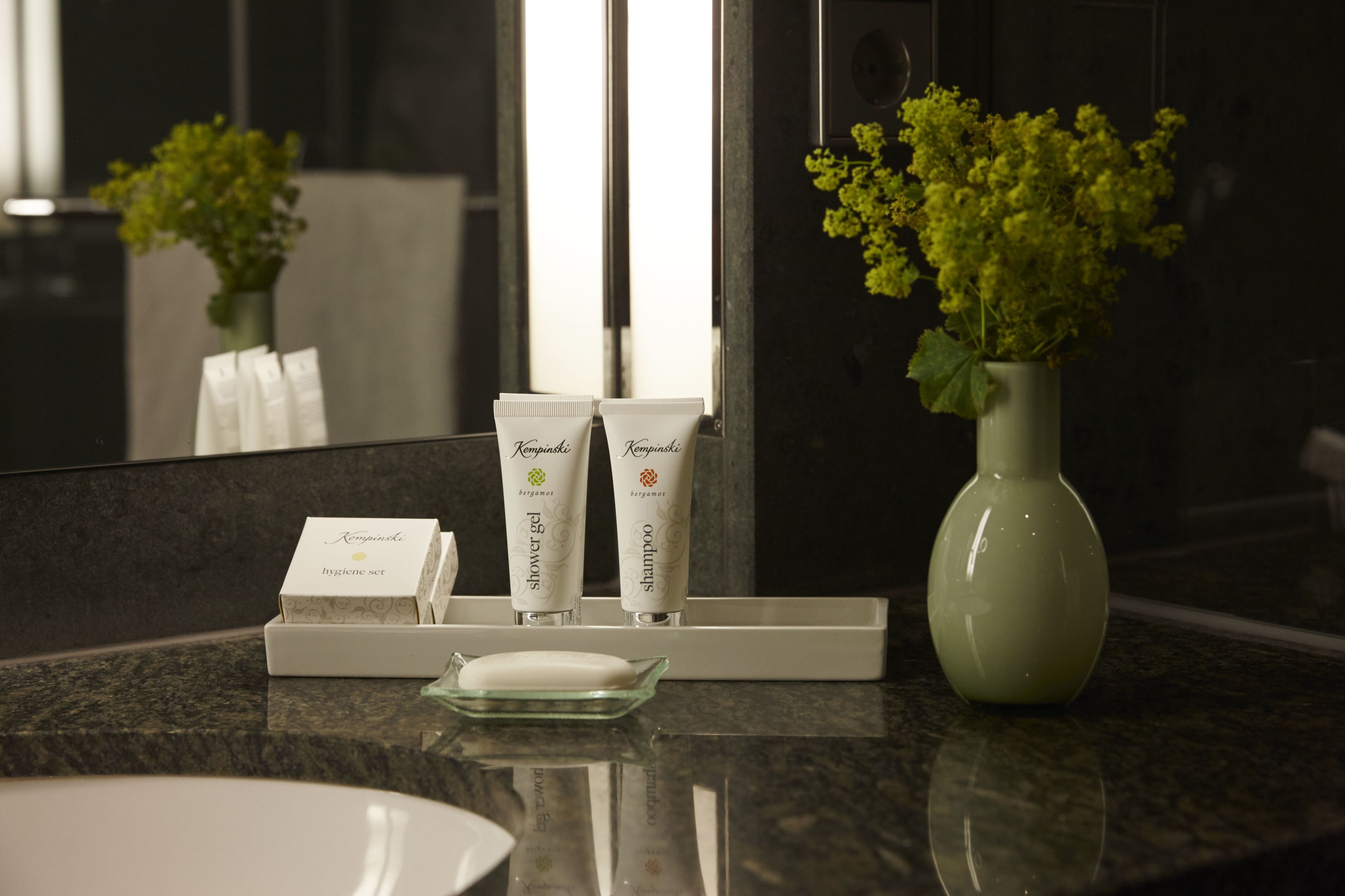 Ein Waschtisch mit Spiegel, eine grüne Vase mit gelben Blumen und Toilettenartikeln, darunter ein Hygiene-Set, Duschgel und Shampoo.