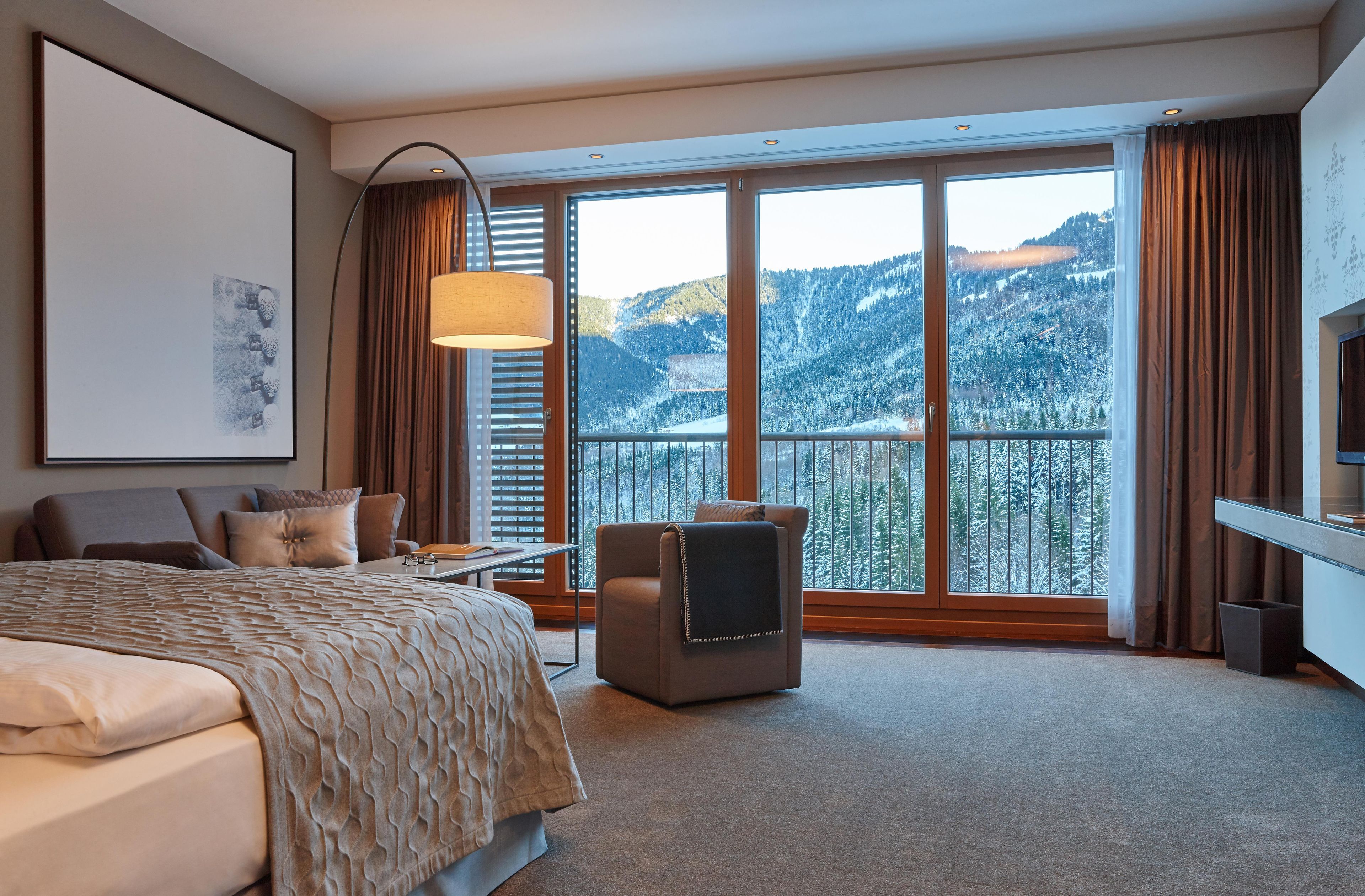 A hotel room with a bed, a desk, a chair, a TV, and a large window with a mountain view.