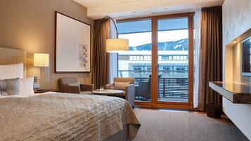 Ein Hotelzimmer mit einem großen Bett, einem Schreibtisch, einem Stuhl, einem Fernseher und einem Balkon mit Blick auf ein Gebäude und Berge.