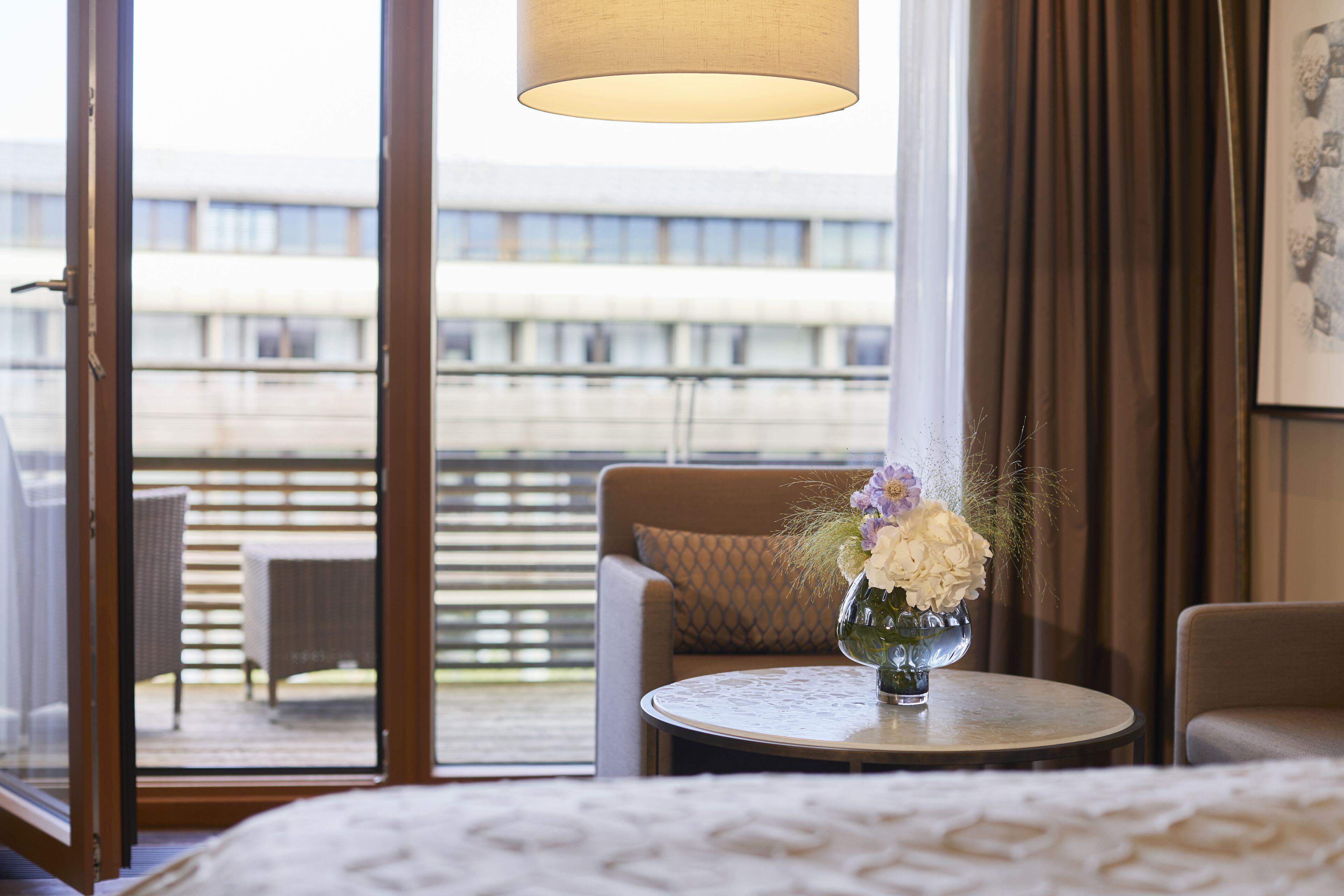 A hotel room with a bed, a round table with a vase of flowers, a window with a view of a building, and a balcony with outdoor furniture.