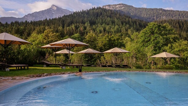 Ein Poolbereich mit Liegestühlen und Sonnenschirmen, umgeben von einem Wald und Bergen im Hintergrund.