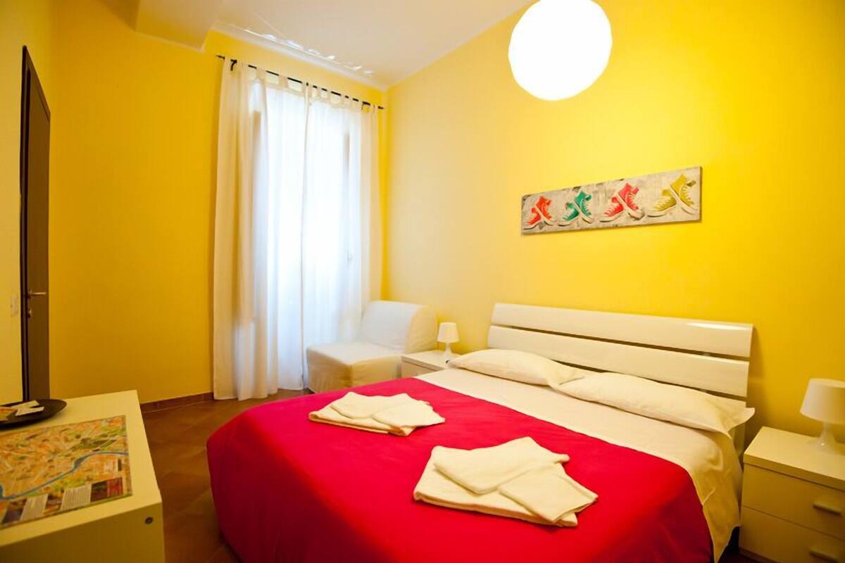 Ein Hotelzimmer mit roter Bettdecke, weißer Bettwäsche, einem Flachbildfernseher, einer Karte auf einem Nachttisch und einem Bild an der Wand.