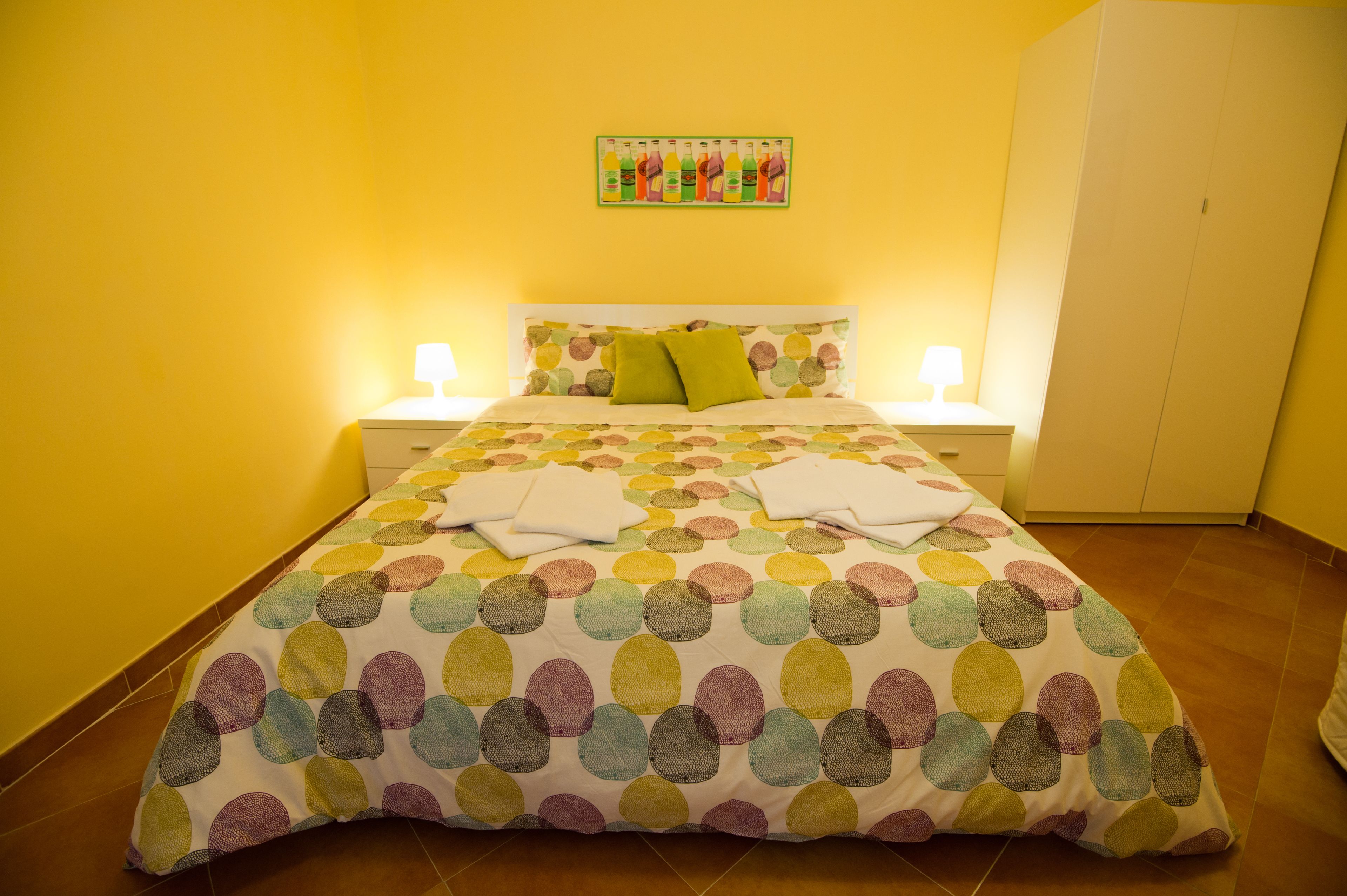 Un lit recouvert d’une courtepointe aux motifs circulaires et colorés, deux oreillers, une table de chevet avec une lampe et un tableau représentant des bouteilles, accroché au mur.
