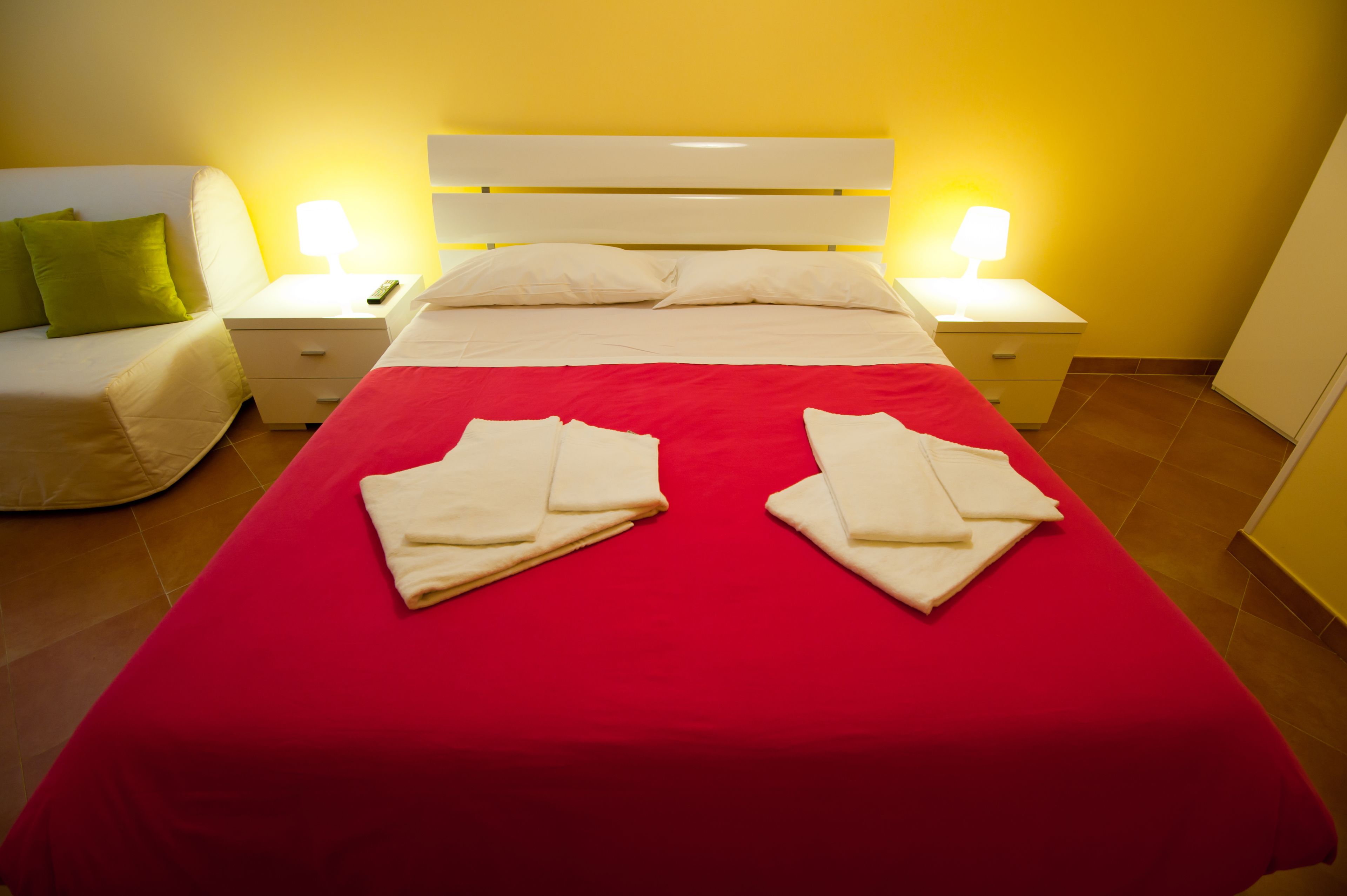 Una habitación de hotel con una colcha roja, cabecero blanco, dos mesitas de noche con lámparas, un sofá blanco con un cojín verde y una alfombra roja.