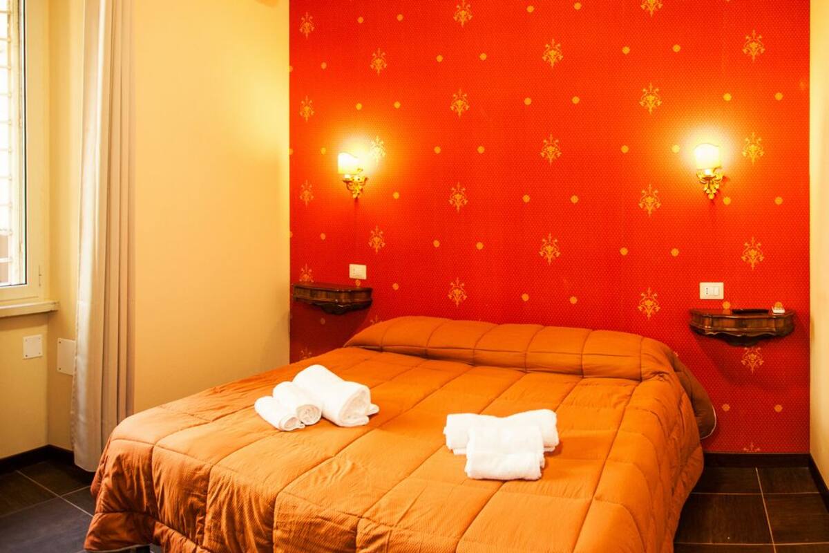 A hotel room with a red patterned wall, two wall-mounted lamps, an orange bedspread, and two neatly folded towels on the bed.