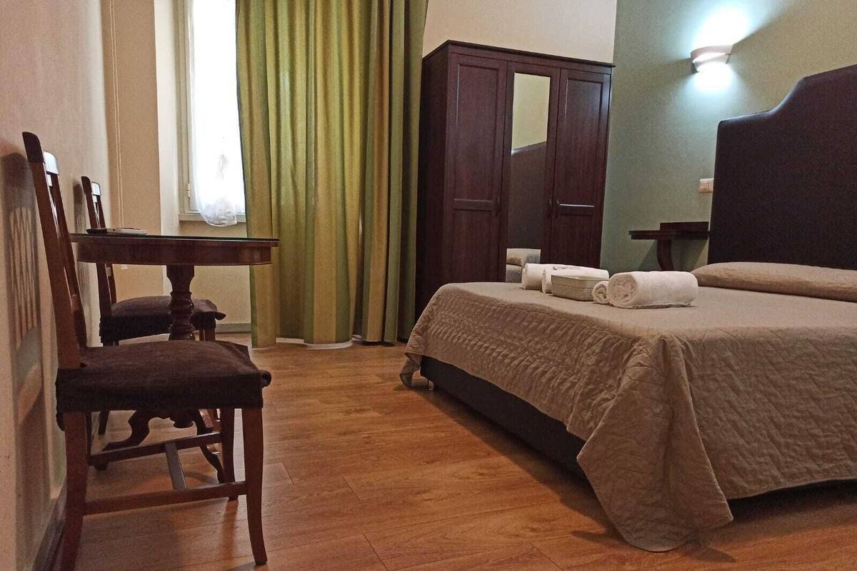 A hotel room with a wooden floor, a bed with a beige bedspread, a wooden chair, a wooden wardrobe, and a window with green curtains.