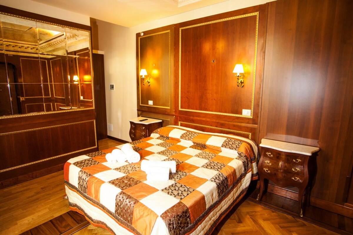 A hotel room with a large bed, wooden paneling, a mirror, two wall-mounted lamps, and a small wooden nightstand.