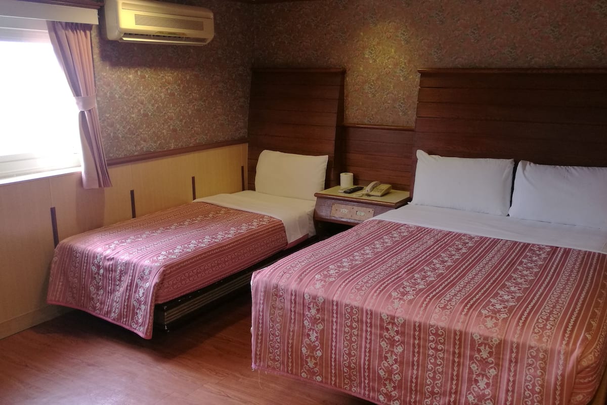 A hotel room with two beds, each with a patterned bedspread, a wooden headboard, a bedside table with a phone and a lamp, and a window with curtains.