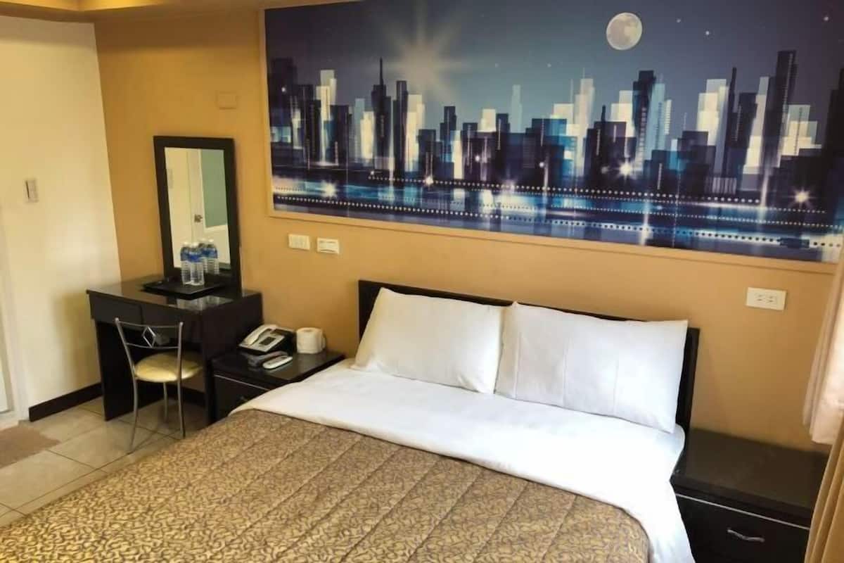 A hotel room with a bed, a desk with a chair, a telephone, and a painting of a city skyline on the wall.