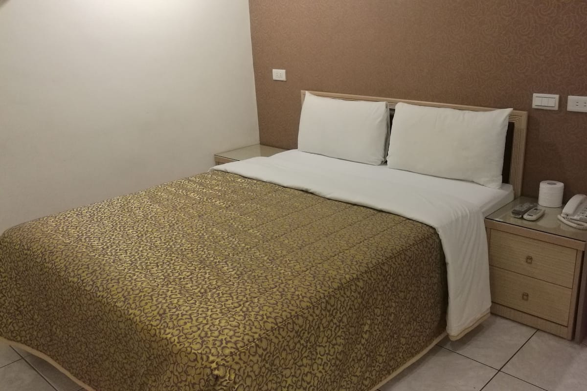 A hotel room with a bed, a bedside table with a phone and a lamp, a small desk with a lamp, and a trash can.