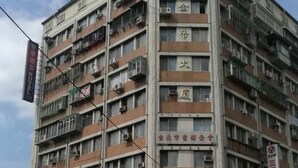 Un edificio de varios pisos con balcones, un letrero con caracteres chinos y una calle con vehículos y peatones.