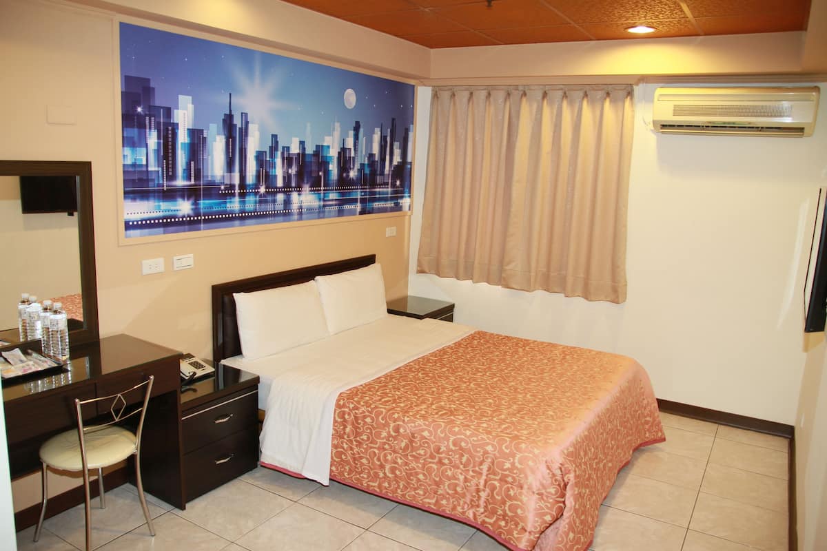 A hotel room with a bed, a desk with a chair, a nightstand, a mirror, and a cityscape painting on the wall.
