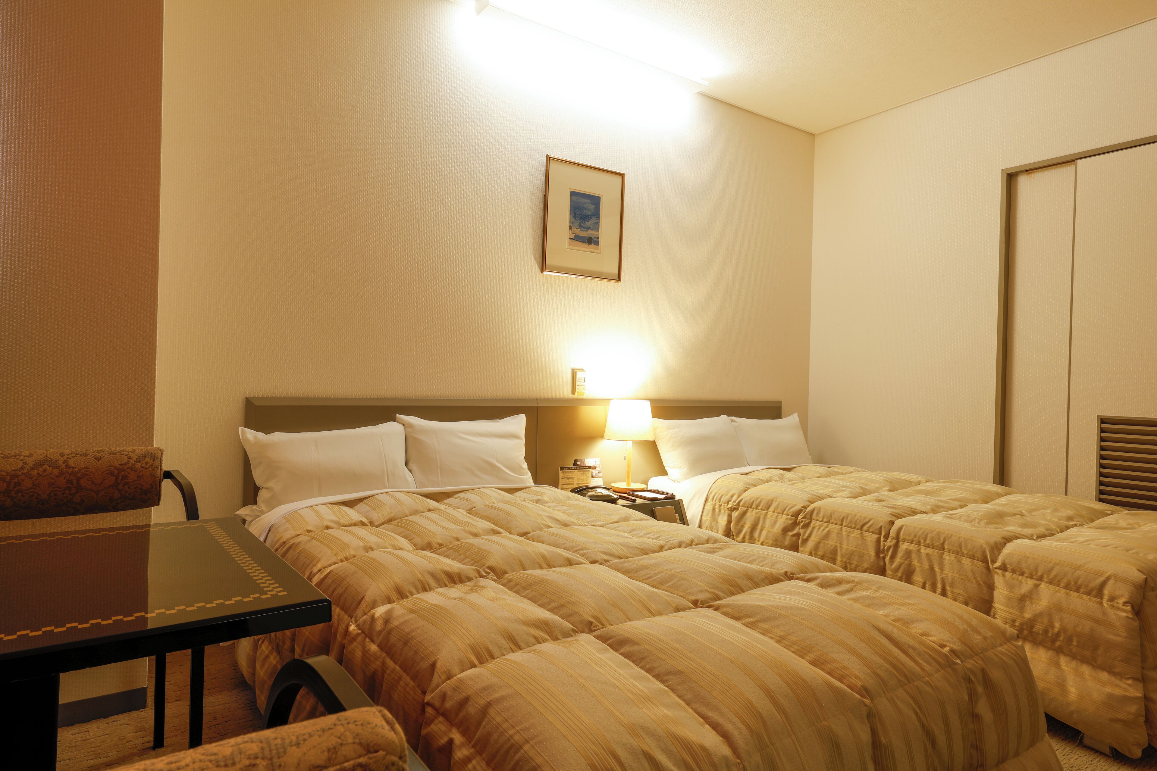 A hotel room with two beds, a desk, a chair, a nightstand with a lamp, and a framed picture on the wall.