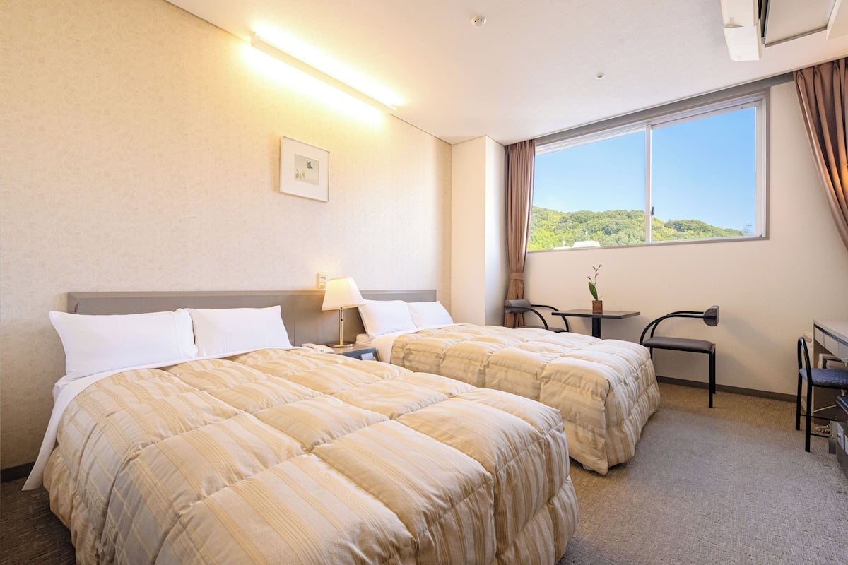 Ein Hotelzimmer mit zwei Betten, einem Schreibtisch, einem Stuhl, einem Fenster mit Blick auf Grünflächen und einer Deckenleuchte.